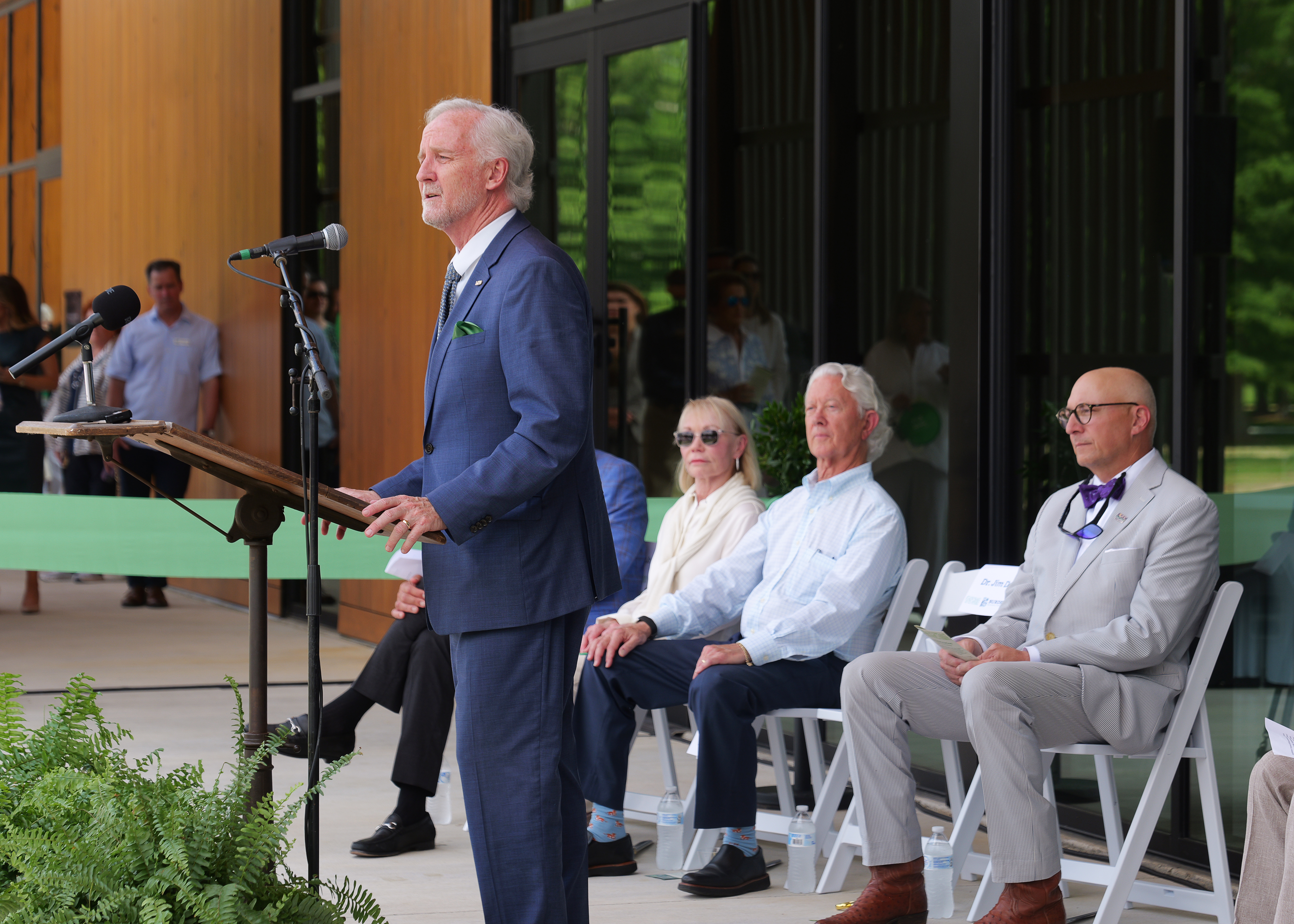 Man speaking at lectern with people seated nearby.